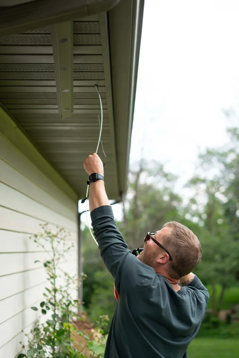 A Lumenary Pro installer reaching up to route wiring into the soffit of a home during a daytime installation.
